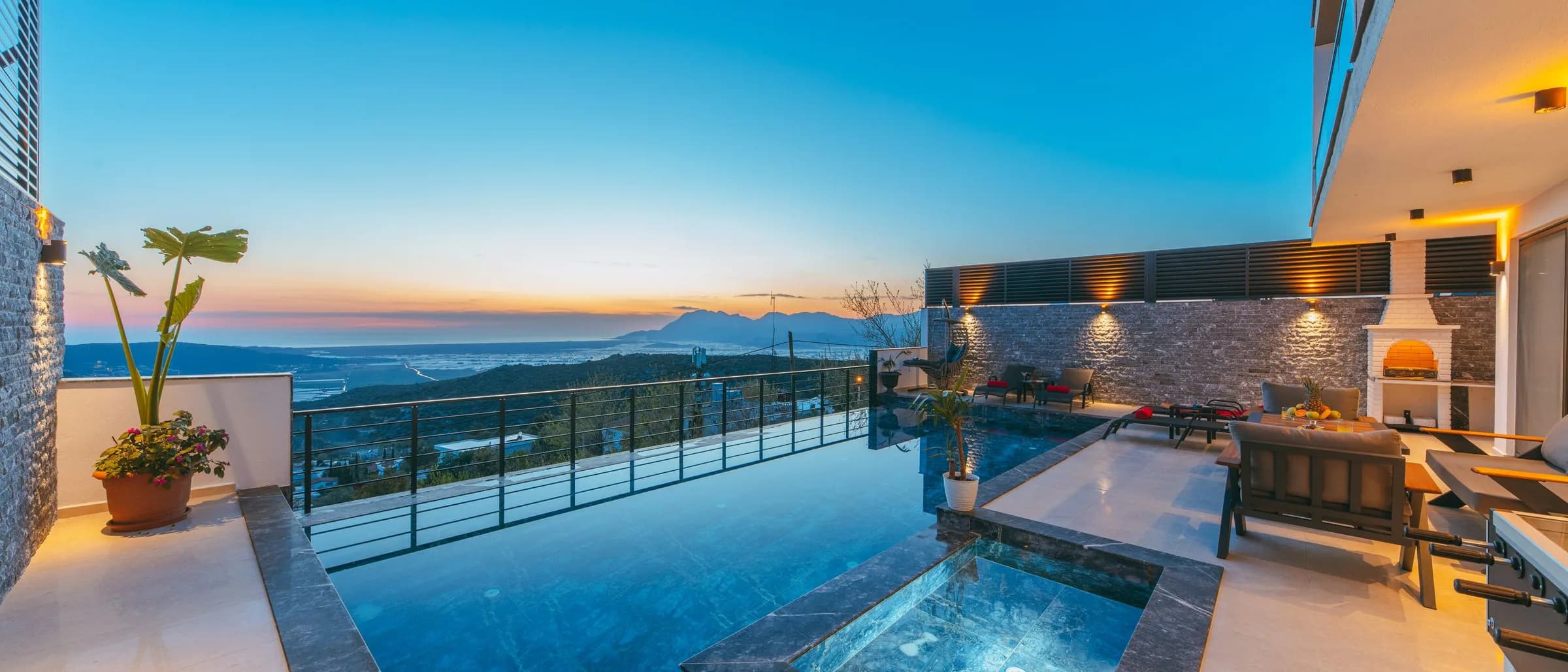 Luxury terrace infinity pool and spa at dusk with mountain valley views and warm patio light